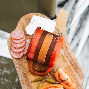 Rustic enamel mug with premium leather sleeve on wooden surface, outdoor camping style, top view