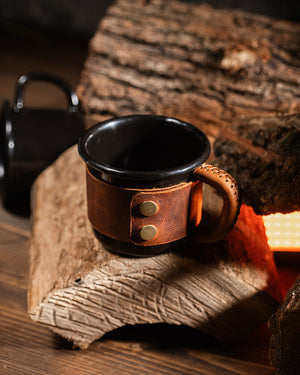 Metal enamel mug with leather sleeve secured by dual brass snap closures, stainless hardware detail
